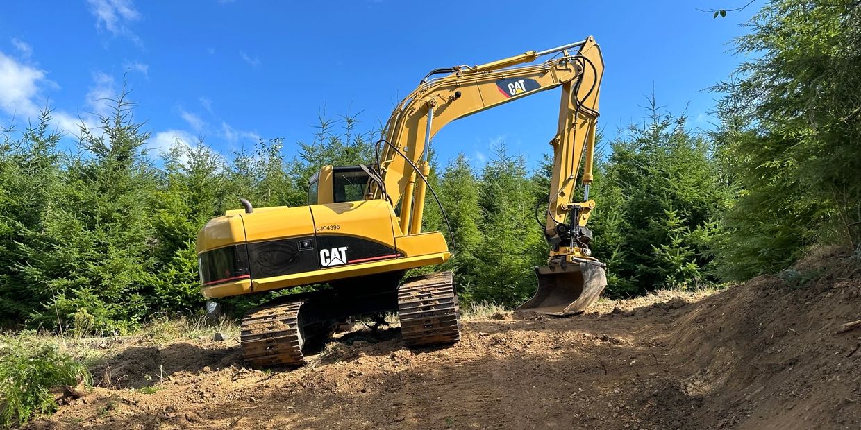 Yellow CAT excavator working on a dirt path surrounded by evergreen trees.