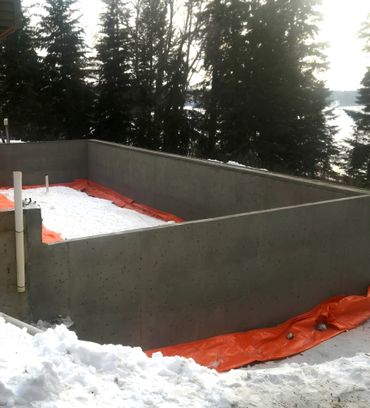 Concrete foundation walls with snow and orange tarp outside in a wooded area.