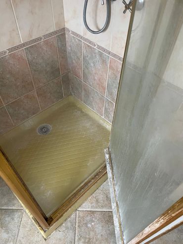 Close-up of a worn shower base with tiled walls and a glass door.