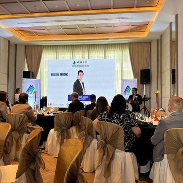 Guests seated at a formal event with a speaker and presentation screen in the background.