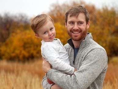 A happy man holding a playful toddler outdoors with autumn foliage in the background.
