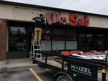 A worker installs a 'Mr. Sub' sign on a storefront using a ladder.