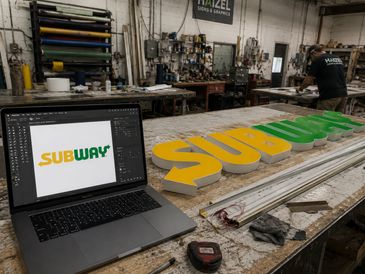 A workshop with a Subway logo design on a laptop and a large 3D Subway sign on the table.