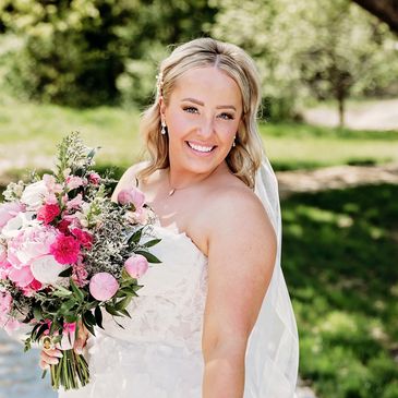 A smiling bride in a white dress holding a vibrant pink and white bouquet outdoors.