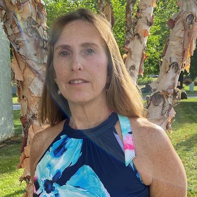 Woman in a blue floral dress standing by a tree on a sunny day.