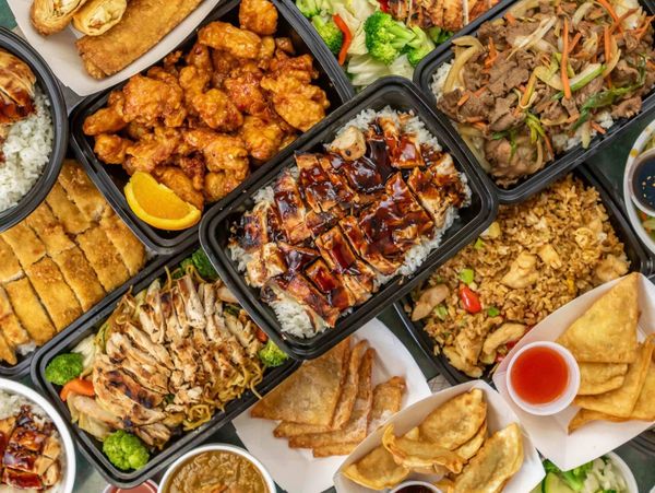 A variety of Asian takeout dishes including rice, grilled chicken, fried dumplings, and spring rolls.