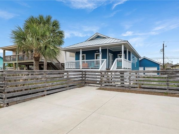 Charming coastal cottage with front porch with fenced yard and palm tree under a bright coastal sky.