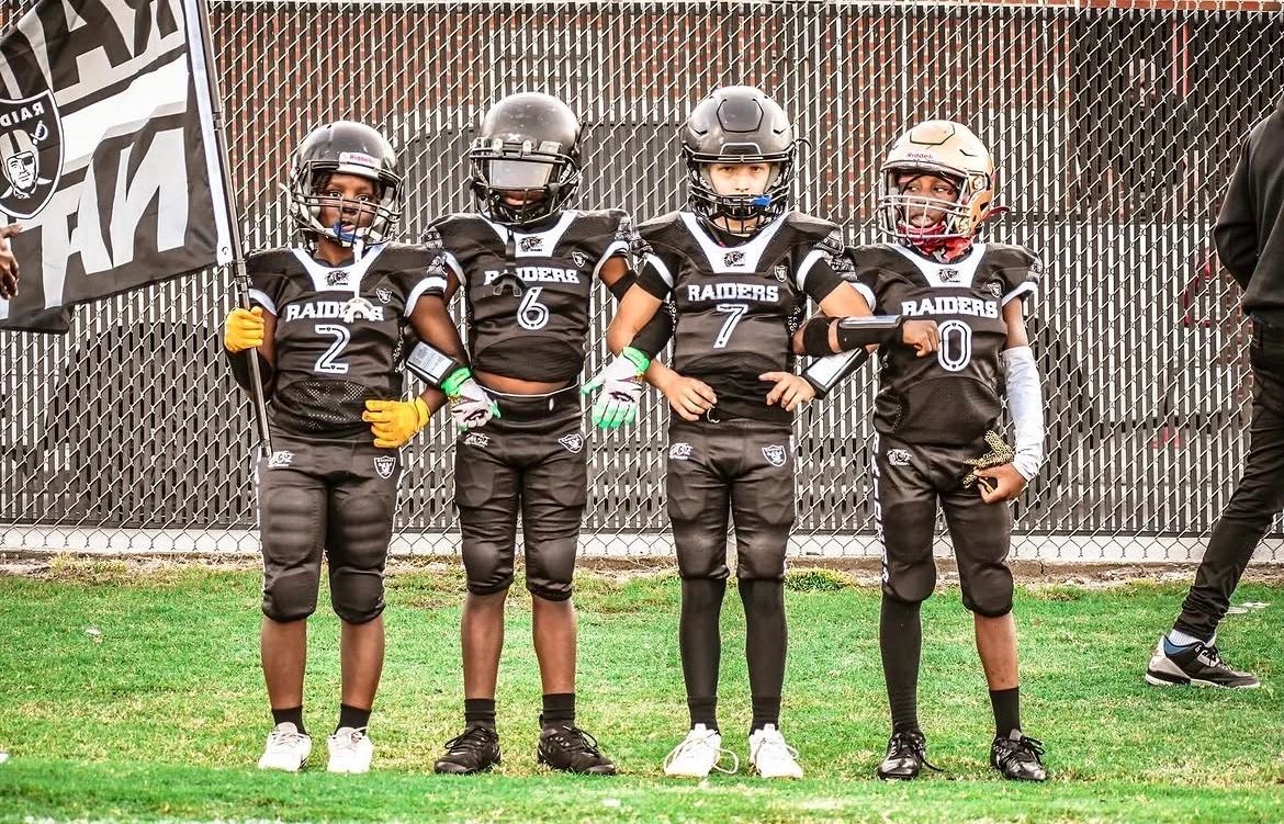Four young football players in Raiders uniforms stand in a line on the field.