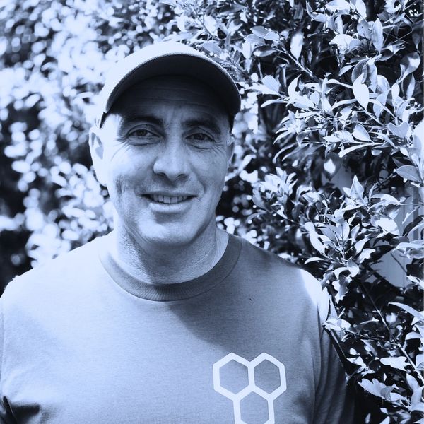 Man wearing a cap and t-shirt standing near leafy bushes, smiling.