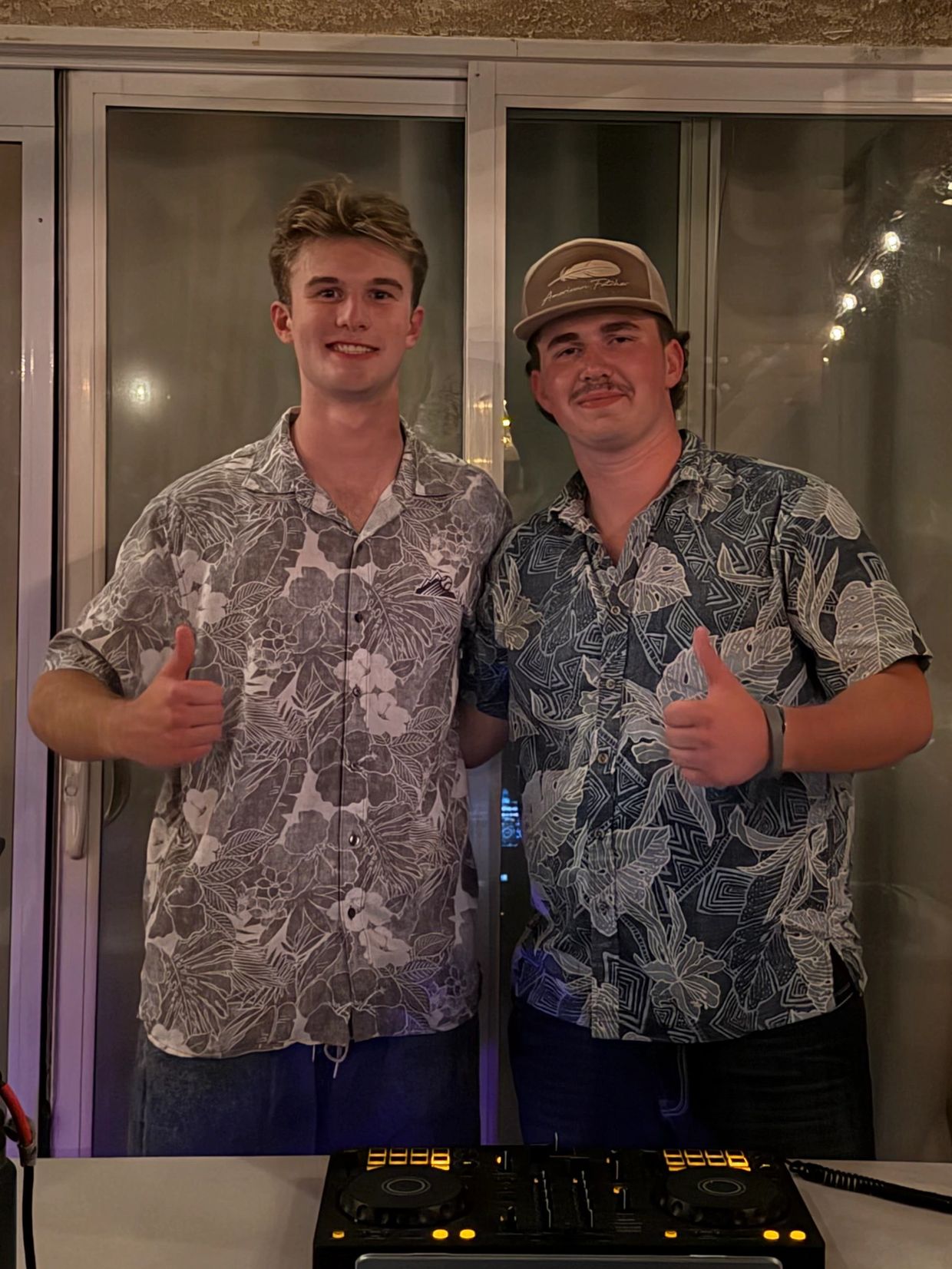 Two young men in floral shirts giving thumbs up, standing behind DJ equipment.