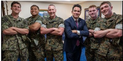 Five men, four in military uniforms and one in a suit, stand with arms crossed indoors.