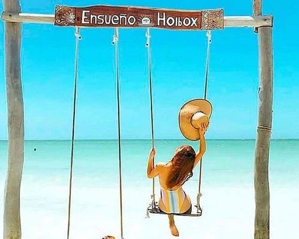 Woman on a swing at a serene beach with a hat, under 'Ensueño Holbox' sign.