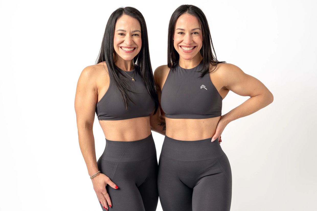 Two fit women in matching gray workout outfits smile at the camera.