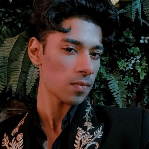 Young man with styled hair wearing a dark embroidered jacket against a leafy background.