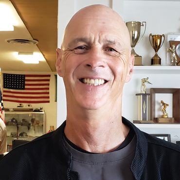 Smiling bald man in front of trophies and an American flag.