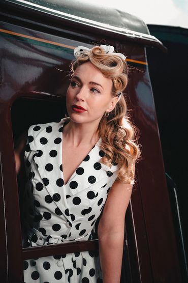 Claudia, leans out of train window. Hair is styled with multiple victory rolls