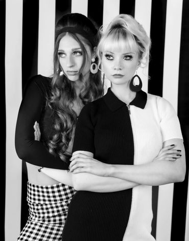 Two women in black and white outfits posing against a striped background.