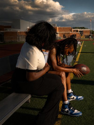 Two people sit on a bench at a sports field, one holding a football.