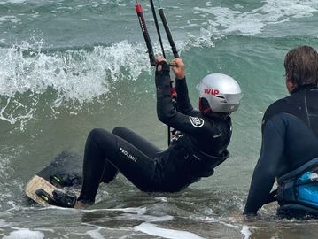 Two people in wetsuits near ocean waves, one preparing to kite surf.
