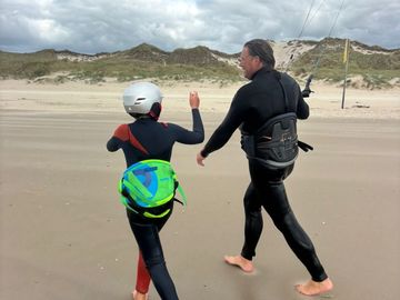 Two people in wetsuits and helmet walking on a beach with kiteboarding gear.