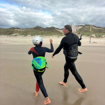 Two people in wetsuits and helmet walking on a beach with kiteboarding gear.