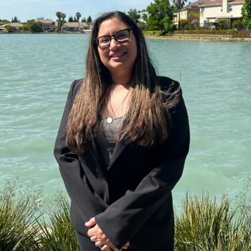 Woman in black blazer smiling by a lakeside on a sunny day.