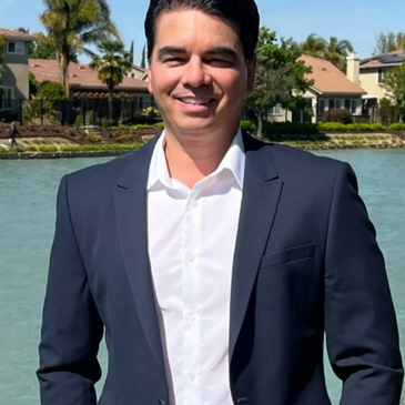 Man in suit standing by a waterfront with houses in the background.
