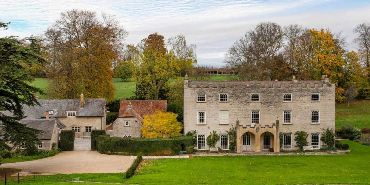 A stately stone manor surrounded by autumn trees and lush green lawns.