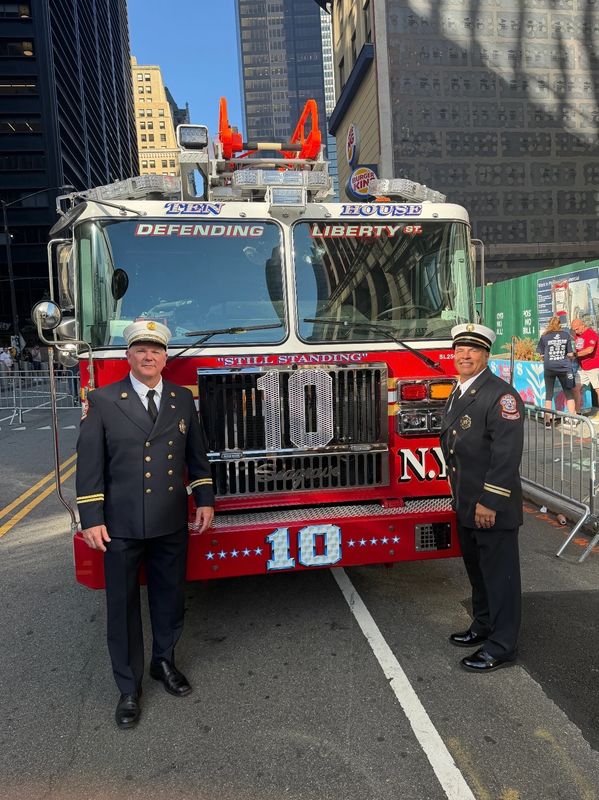 Two firefighters in uniform stand beside a red fire truck in the city.