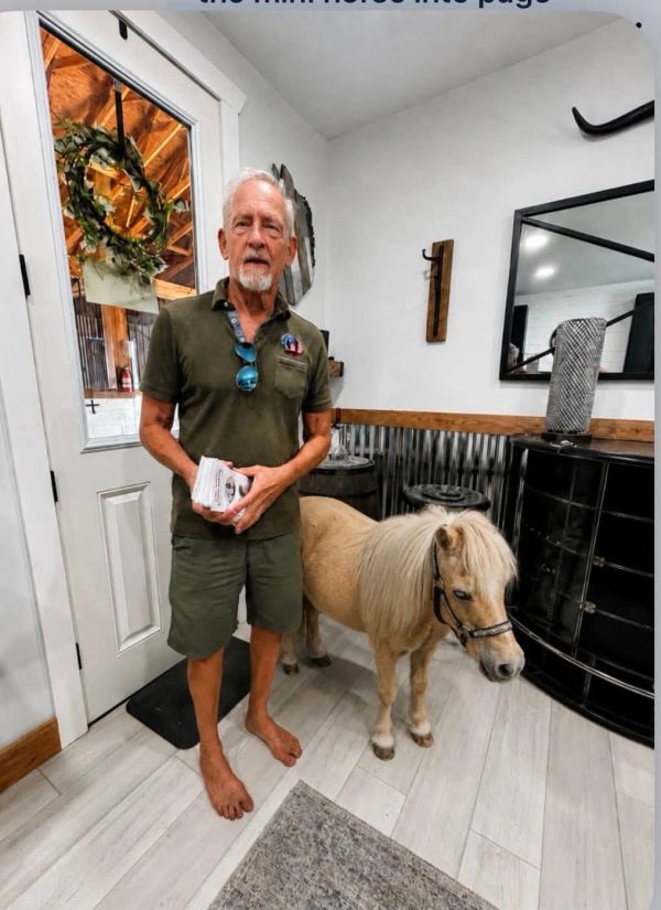 Man standing barefoot with a miniature horse indoors.