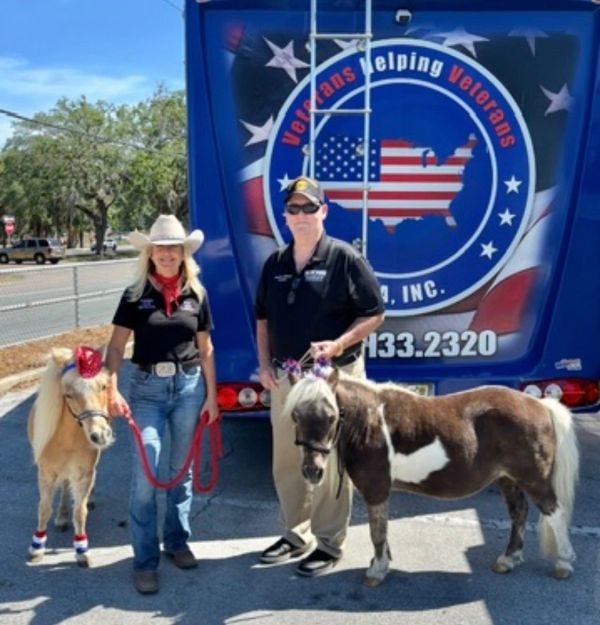 Two people with miniature horses in front of a veterans help truck.
