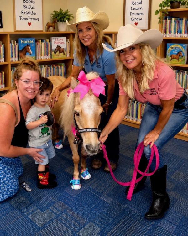 Three women and a child with a miniature horse in a library.