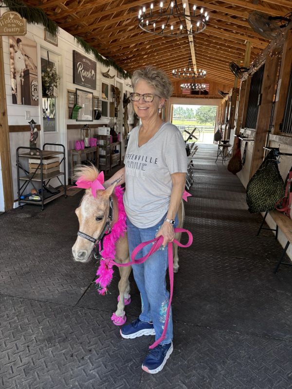 Woman smiling with a decorated miniature horse in a rustic stable.