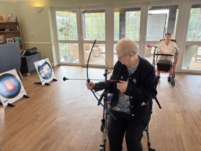 Elderly woman practicing archery indoors with walker support.
