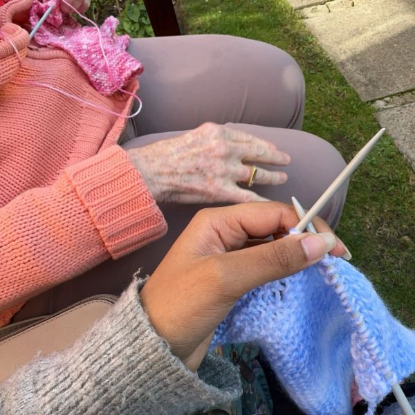 Two people knitting together outdoors, one with pink yarn and the other with blue yarn.