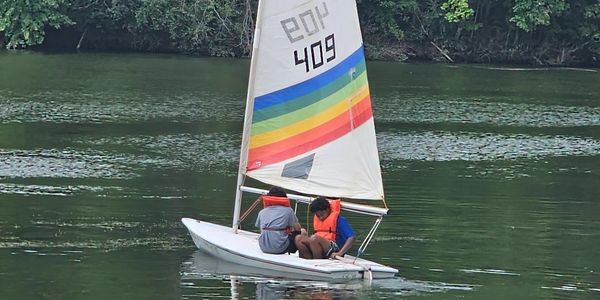 Two boys sail on an FJ sailboat on  a calm lake with trees in the background.
