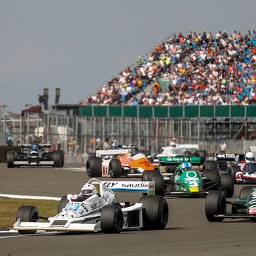 Vintage Formula 1 cars racing on a track with a large crowd in the stands.