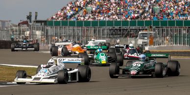 Historic Formula 1 cars racing on track in front of a large crowd.