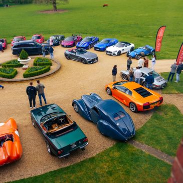 A gathering of luxury and vintage cars on a gravel driveway with people mingling.