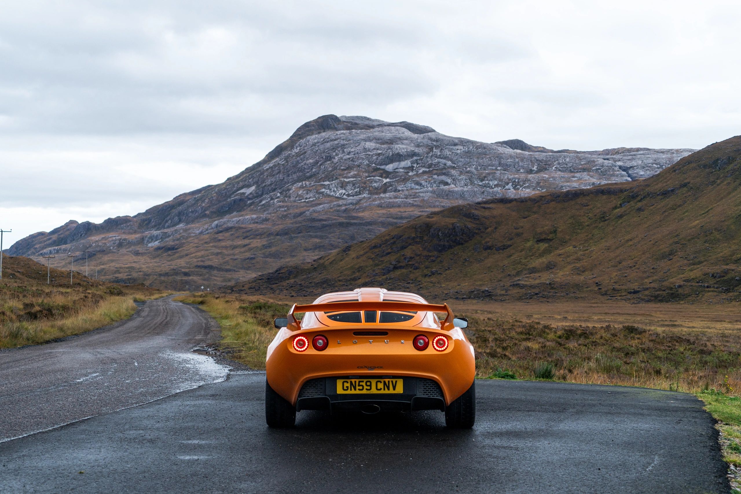 Orange Lotus sports car on a rural road with mountainous backdrop.