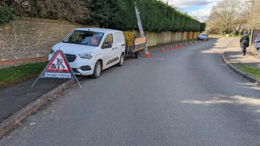 Van with trailer and cones set up for hedge cutting along a suburban street.