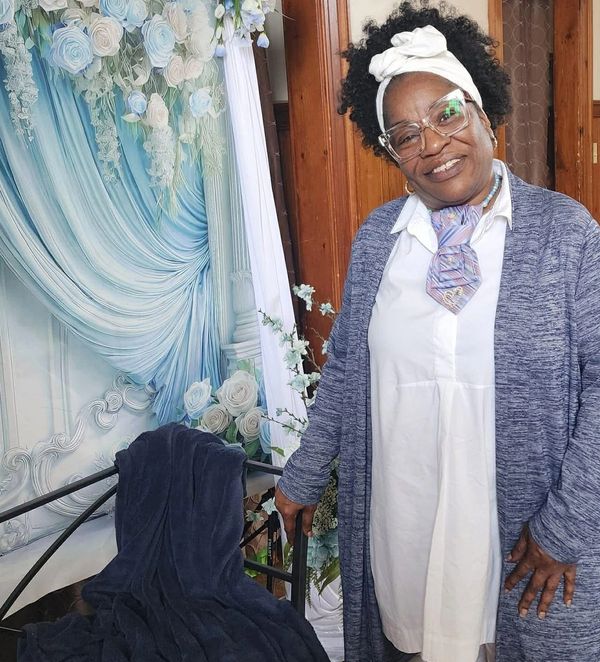 A smiling woman wearing glasses and a headwrap poses indoors near a floral backdrop.