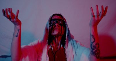 Person with face covered in black paint, hands raised, wearing a white shawl and tattoos visible.