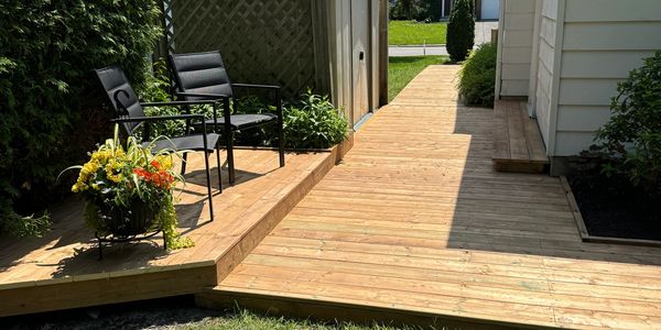 Wooden deck with chairs and flowers beside a white house on a sunny day.