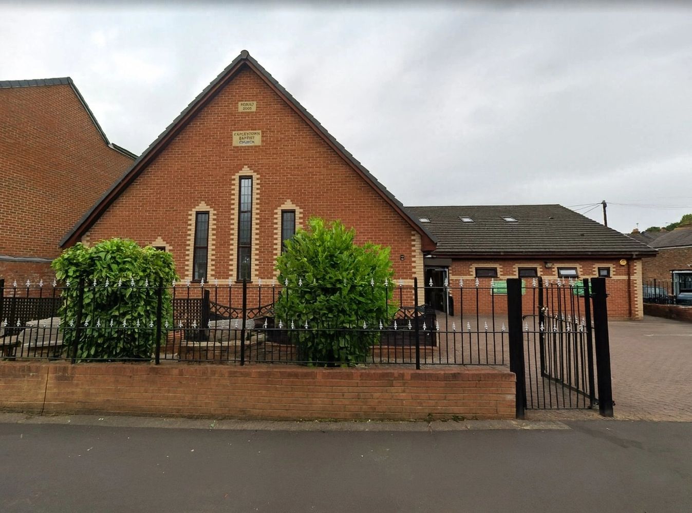 Brick church building with a fenced front garden and paved driveway.