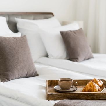 Breakfast tray with coffee and croissants on a cozy bed with brown pillows.