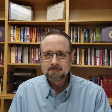 Man with glasses sitting in front of a bookshelf filled with books.