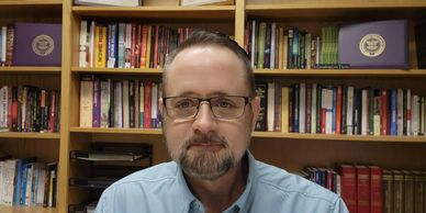 Man with glasses sitting in front of a bookshelf filled with books.