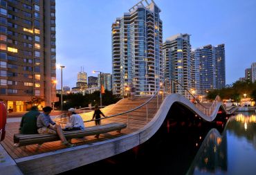 People sitting on a curved wooden bridge in an urban setting at dusk.
