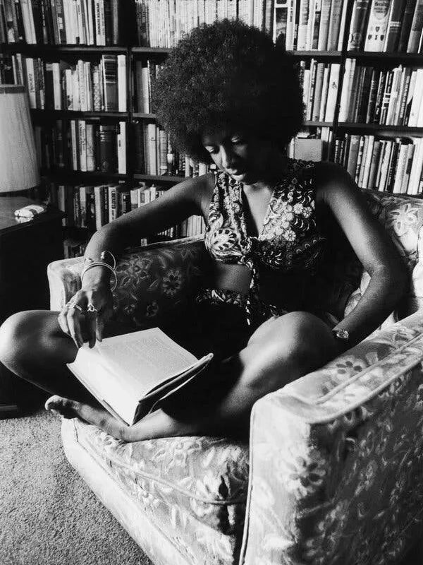 Black woman with afro sitting cross-legged on a chair in front of book shelves reading a book